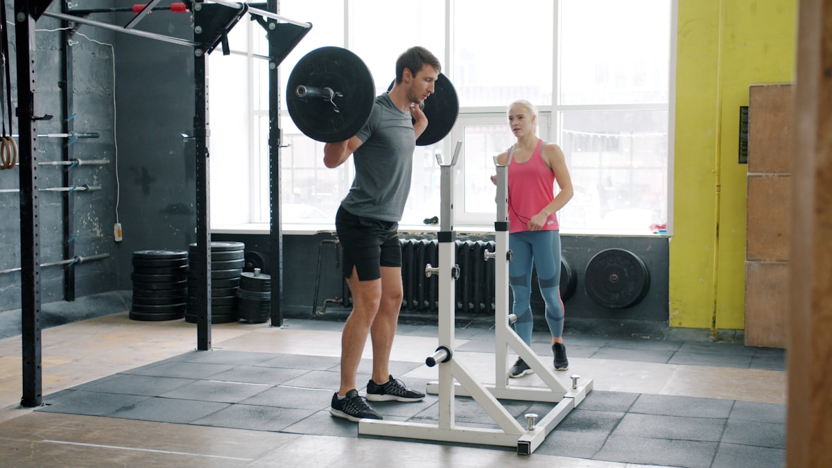Muscular lifter squatting a barbell in a rack with a spotter behind him