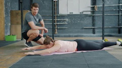 Trainer watches woman exercise on gym floor.