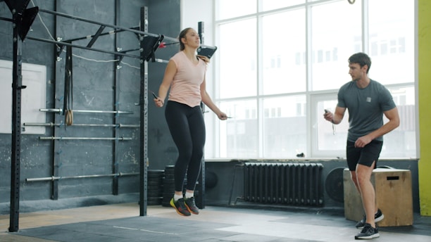 Woman jumps rope while man watches in gym.