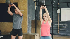Man and woman performing kettlebell swings in gym.