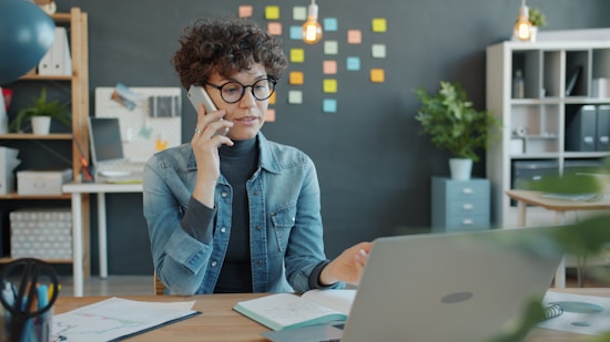 Woman talking on phone at desk with laptop.