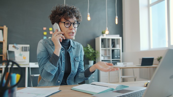 Woman talking on phone at desk with laptop.
