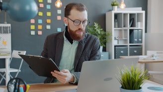 Man with beard working on laptop and clipboard.
