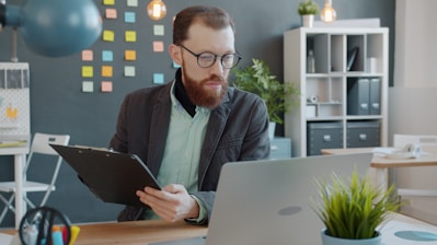 Man with beard working on laptop and clipboard.