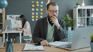 Man thinking at desk with laptop and papers.