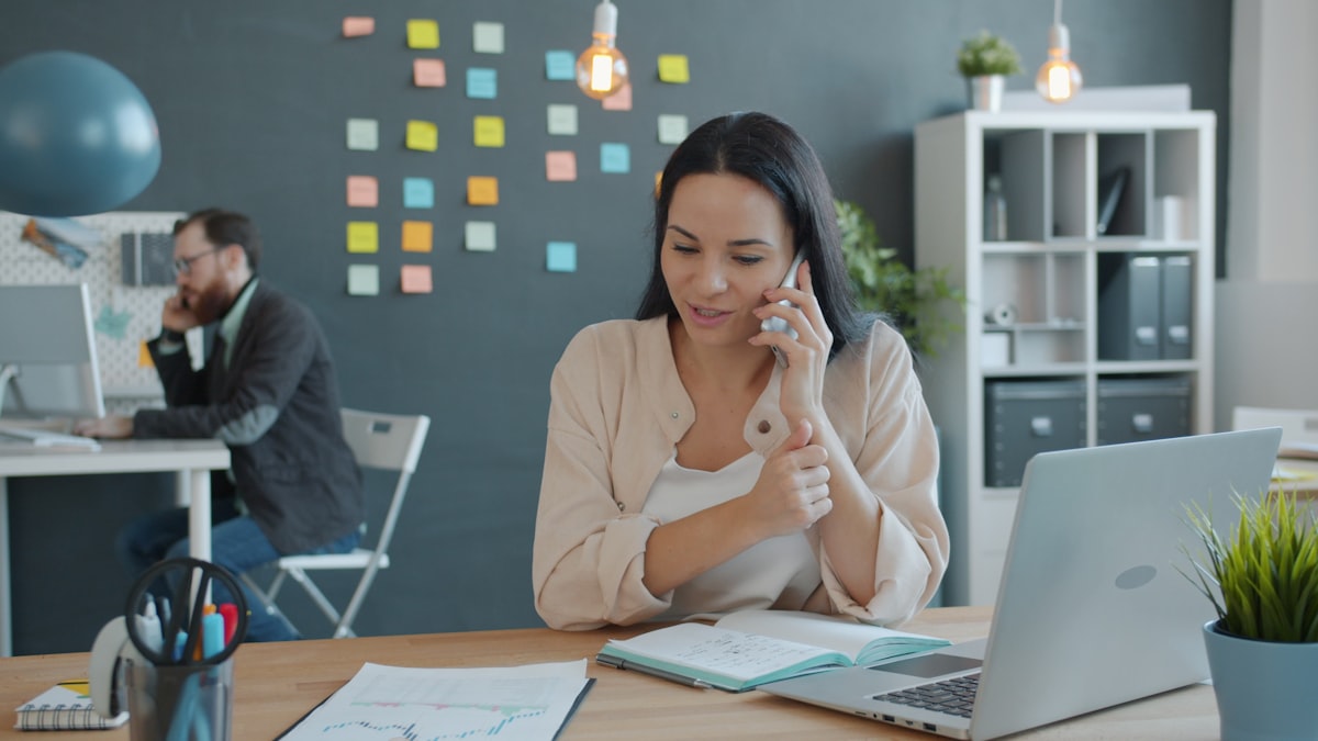 Dirigeante de PME B2B en appel téléphonique au bureau avec ordinateur portable et carnet de notes, scène de prospection commerciale concentrée