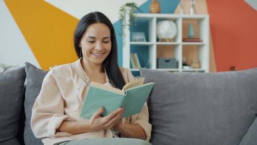 A woman smiles while reading a book on a couch.
