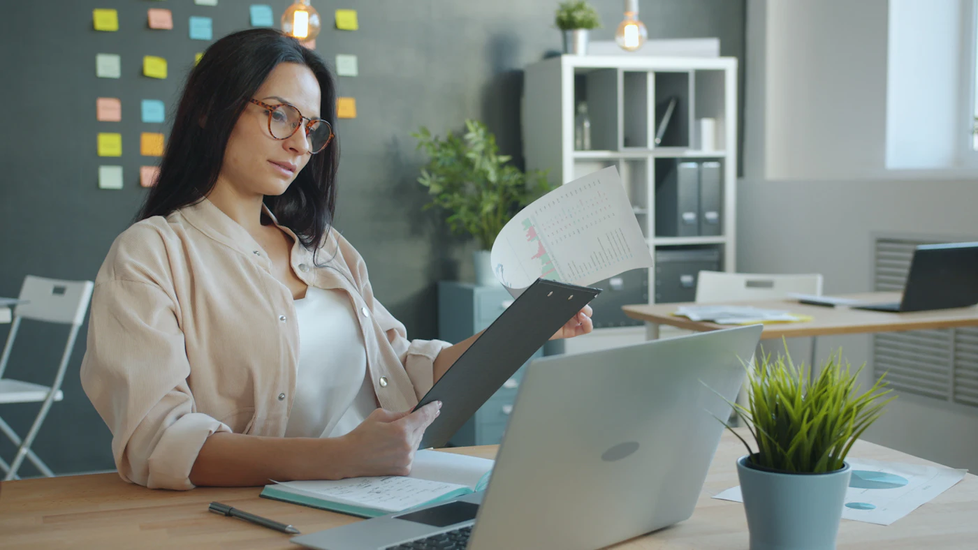 A professional reviewing documents and a laptop at her desk