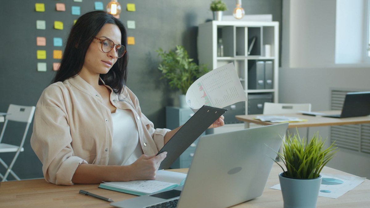 A woman in glasses reviews a document beside her laptop at an office desk
