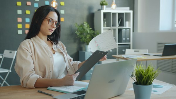 A woman in glasses reviews a document beside her laptop at an office desk