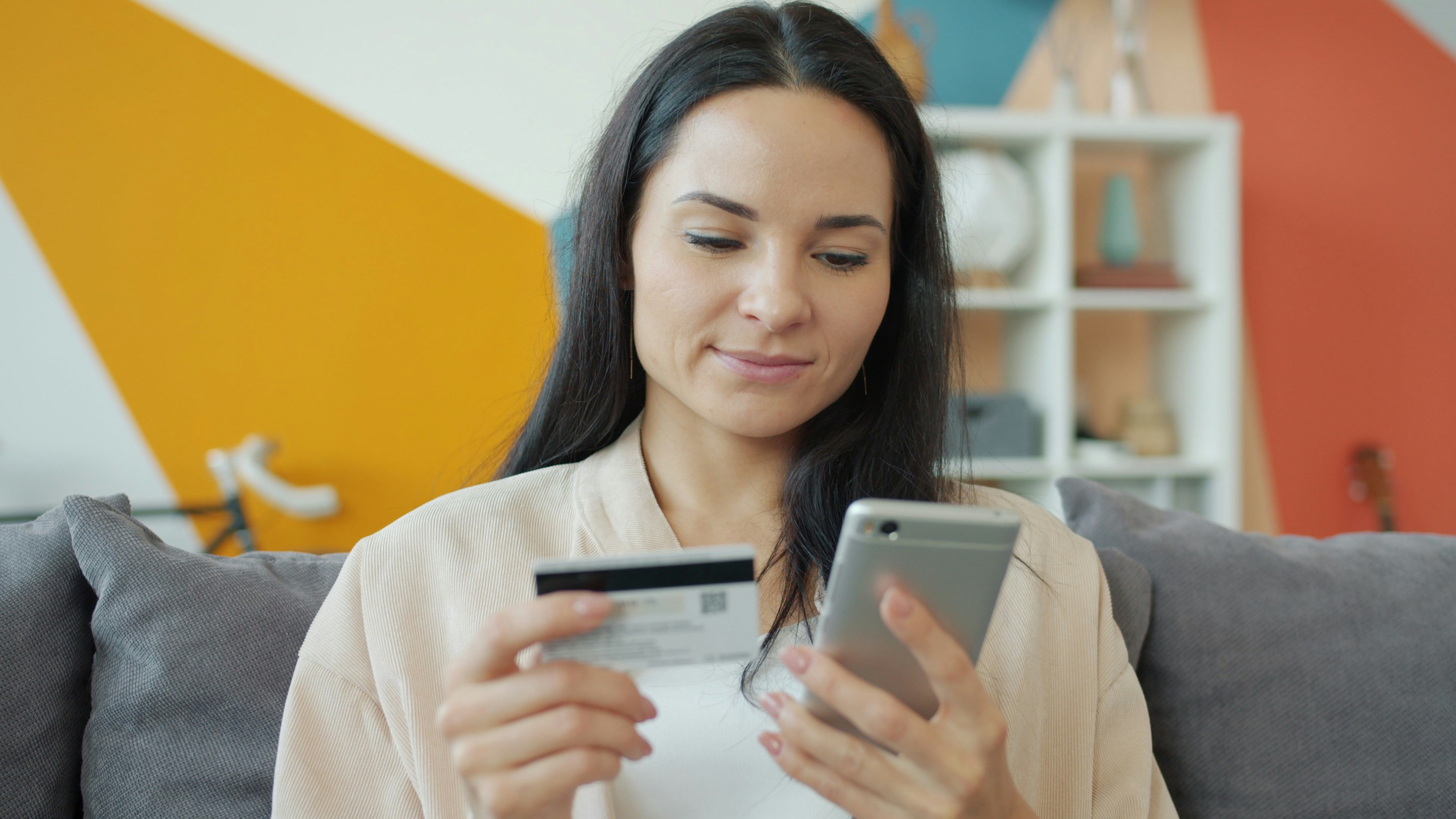 Young woman is making cashless payment online using credit card and smartphone touching screen smiling relaxing on couch in apartment. Technology and youth concept.