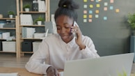 Young woman talking on phone at office desk with laptop.