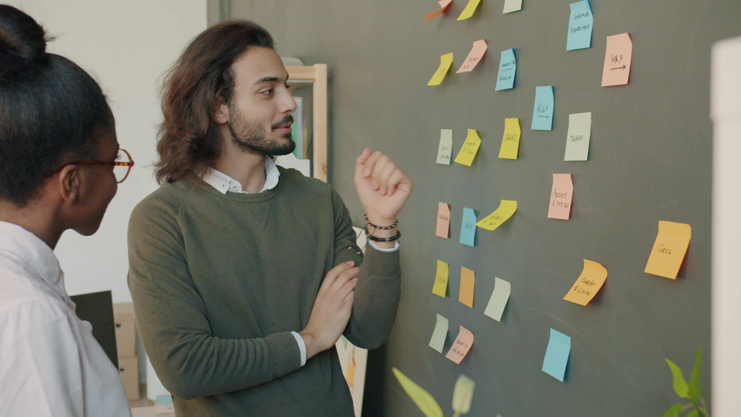 A man in a green jumper and a woman in glasses standing at a grey wall of pinned coloured notes, mid-conversation