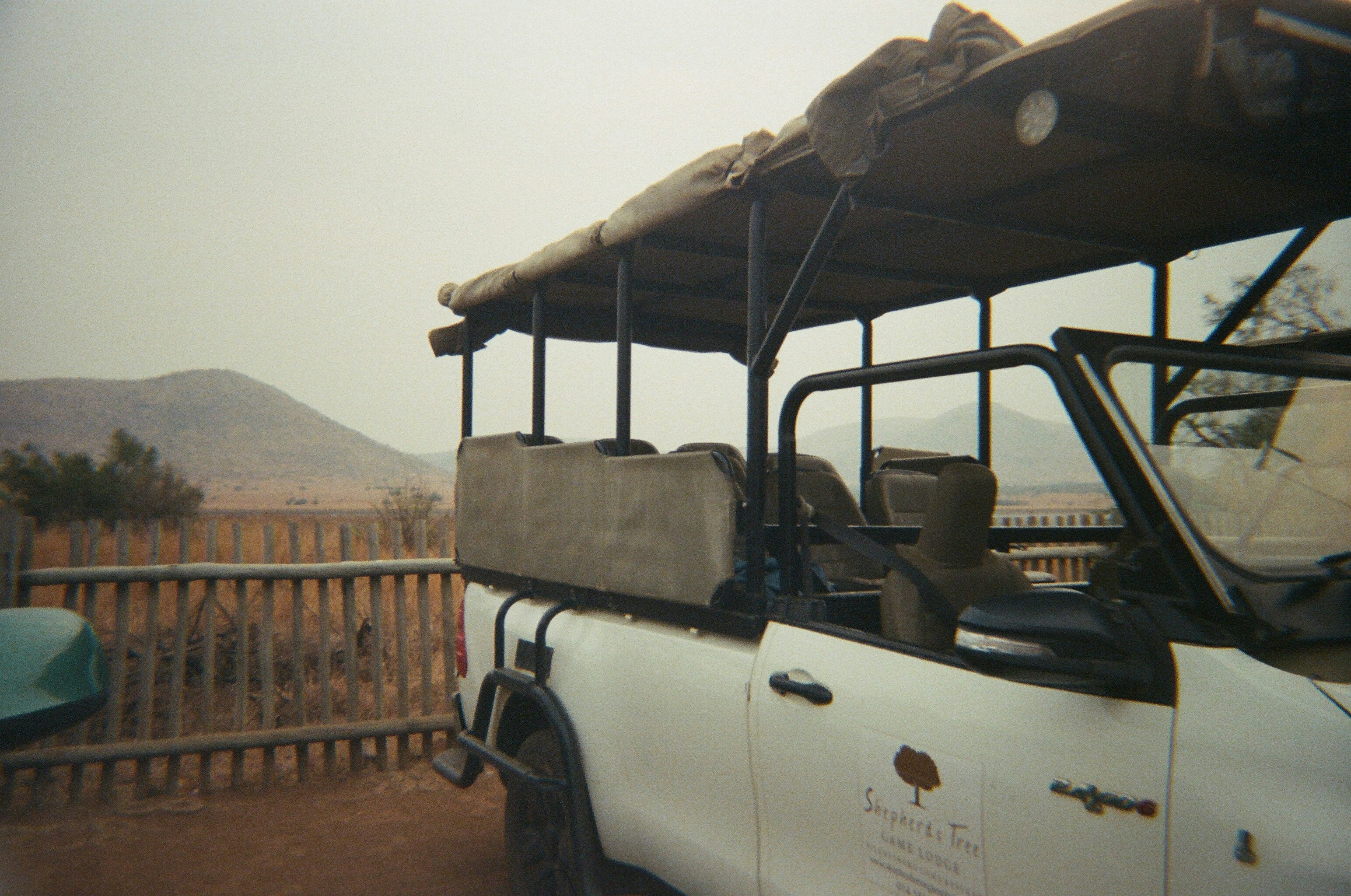 Safari vehicle parked near wooden fence with mountains beyond.