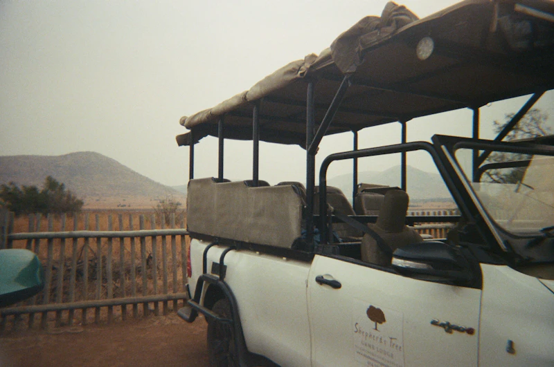 An open safari vehicle parked on level ground beside a lodge fence and walkway, plains and hills beyond