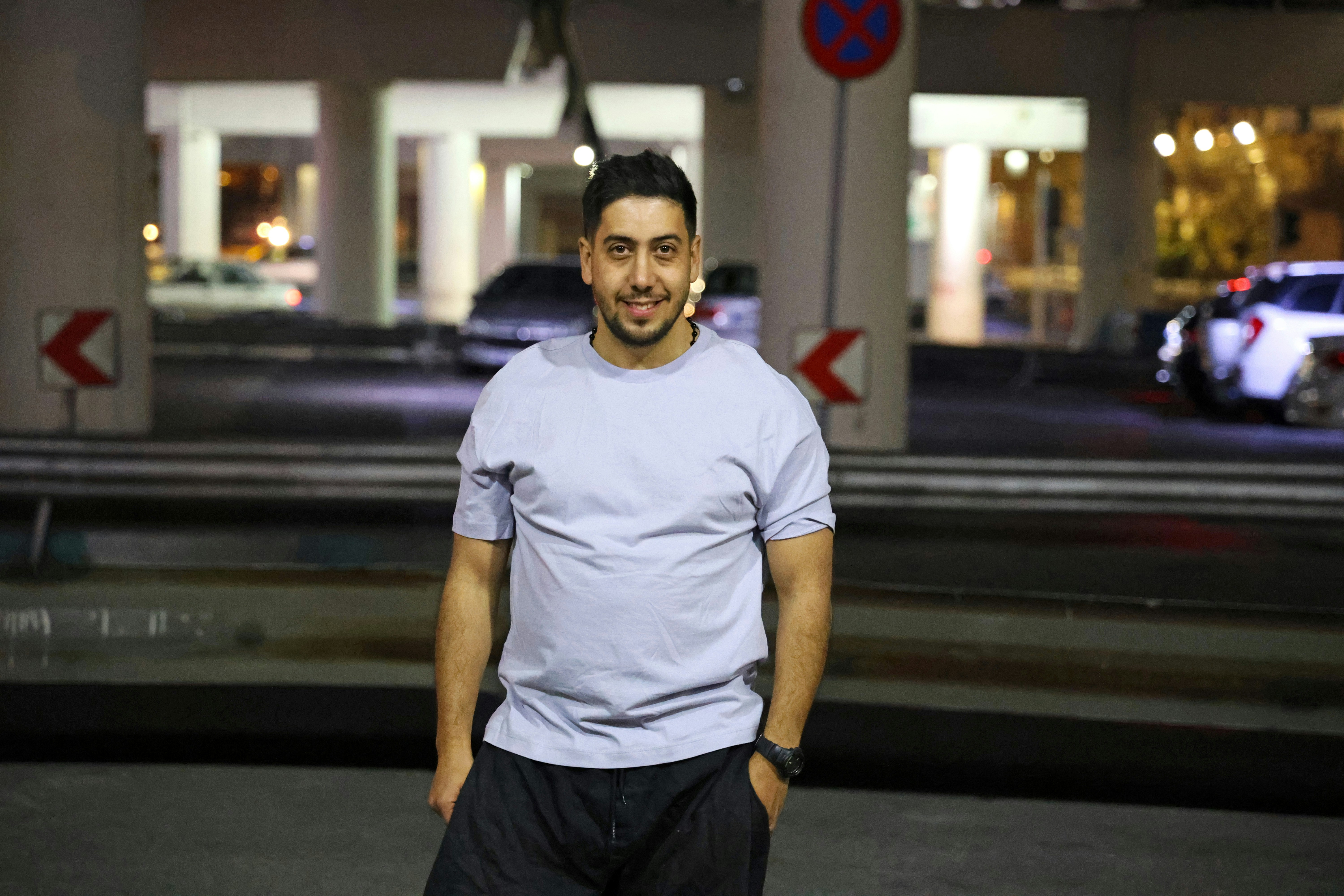 A smiling man stands in a dimly lit urban area.