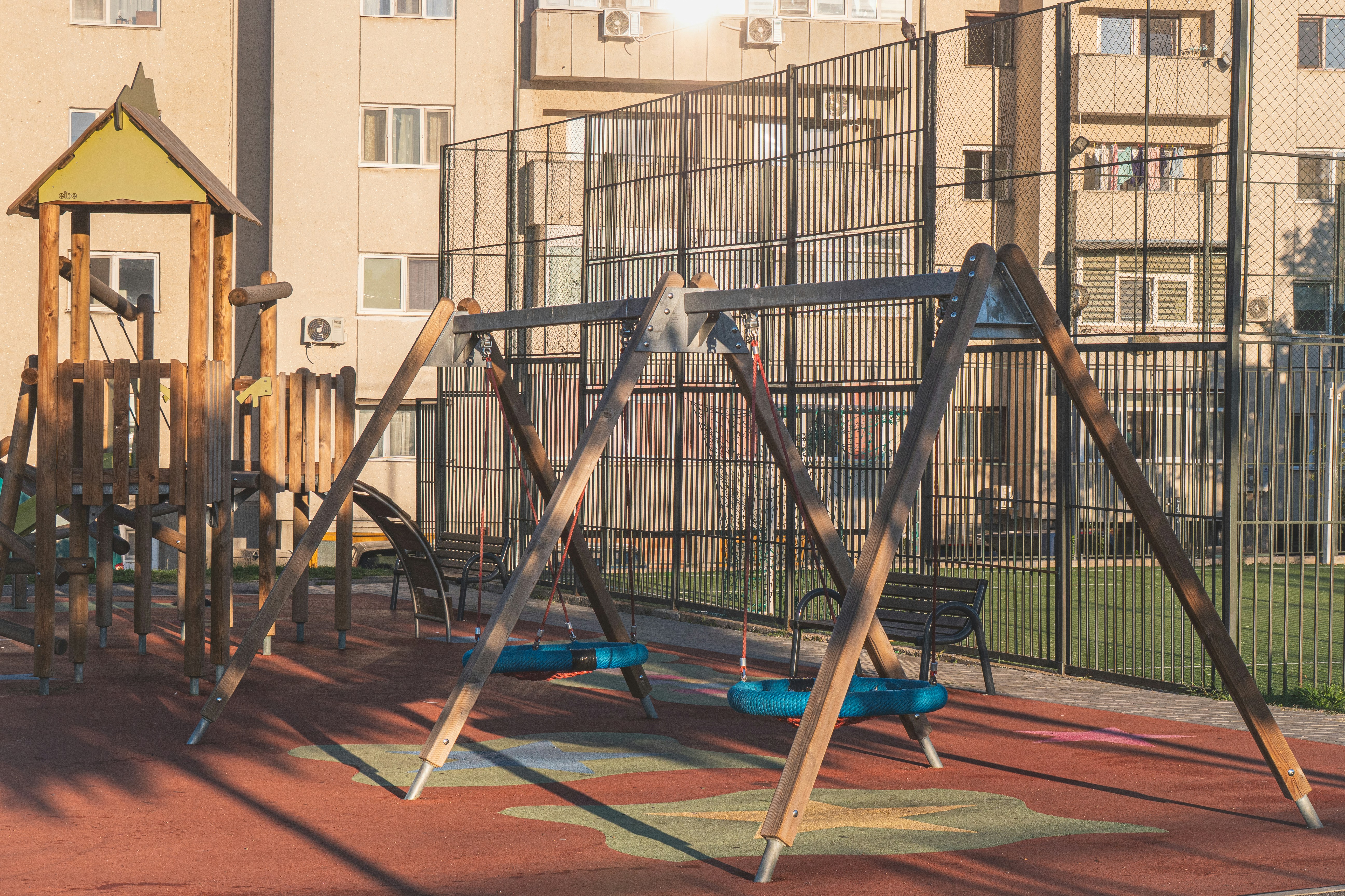 Urban playground scene with no people, photographed in warm, late sunlight. The image focuses on empty swings, geometric structures, and the silent atmosphere of a place usually filled with activity.