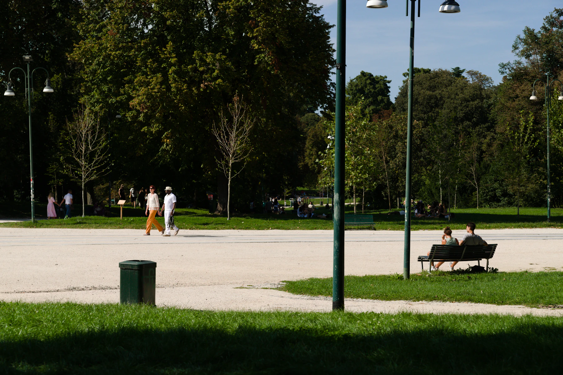 A sunny park with a wide gravel path, two people walking on the left and two people sitting on a bench on the right