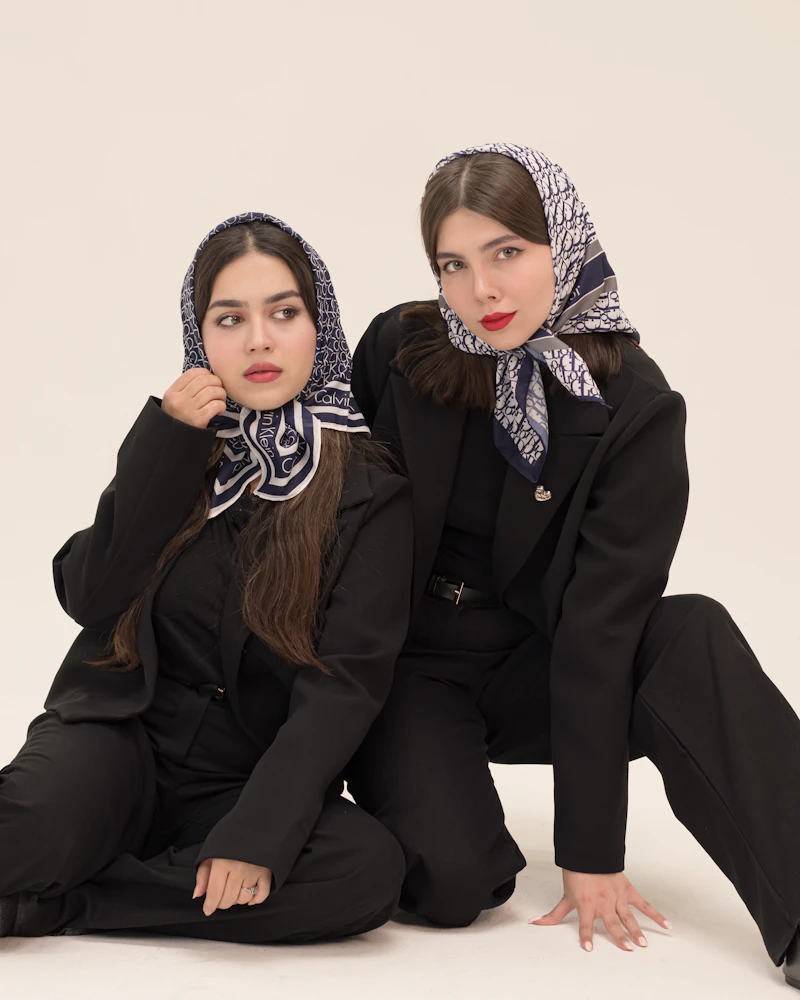 Two women in black suits and patterned headscarves seated on a pale studio floor.