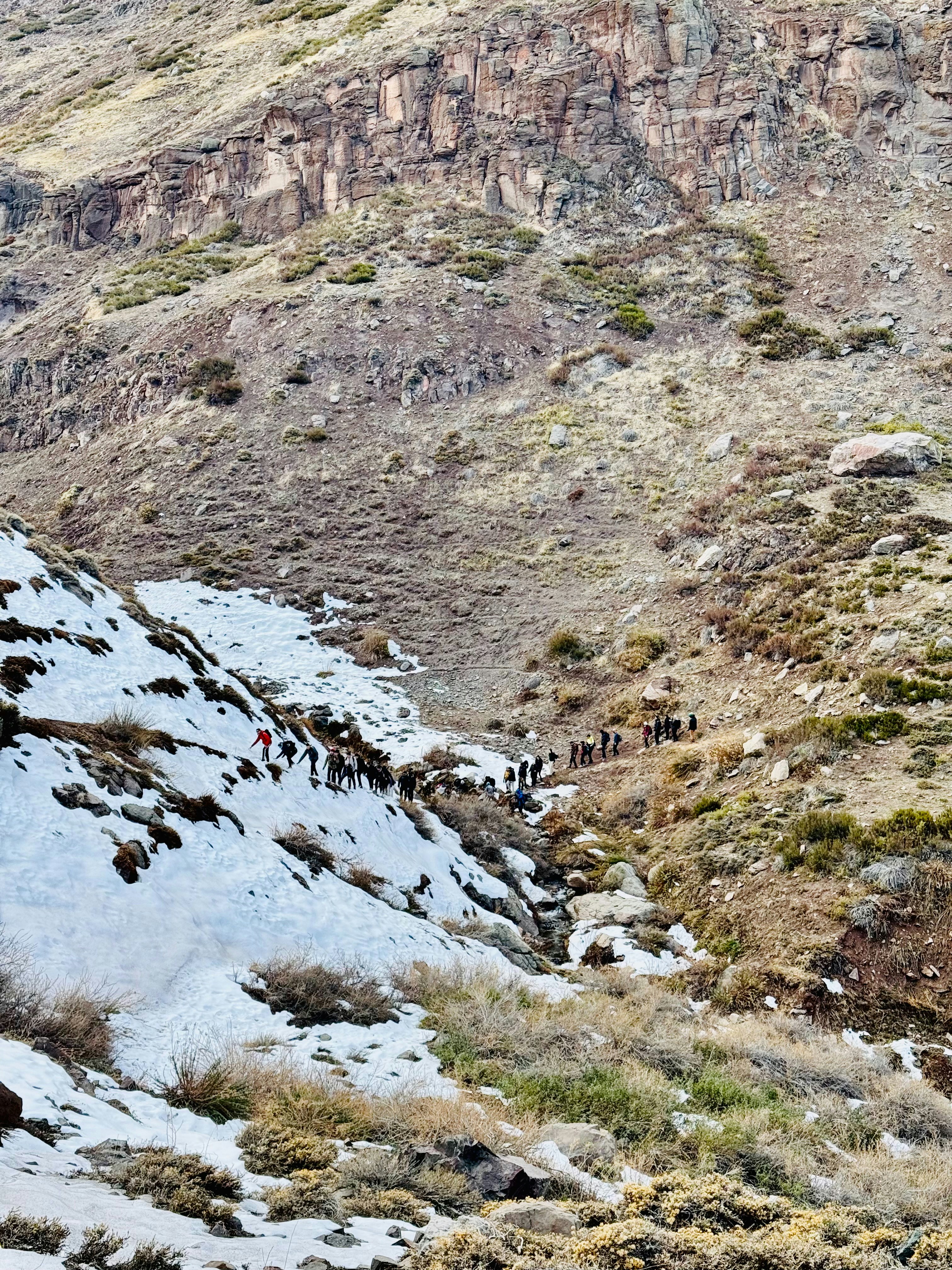 Hikers ascend a snowy mountain path in arid terrain.
