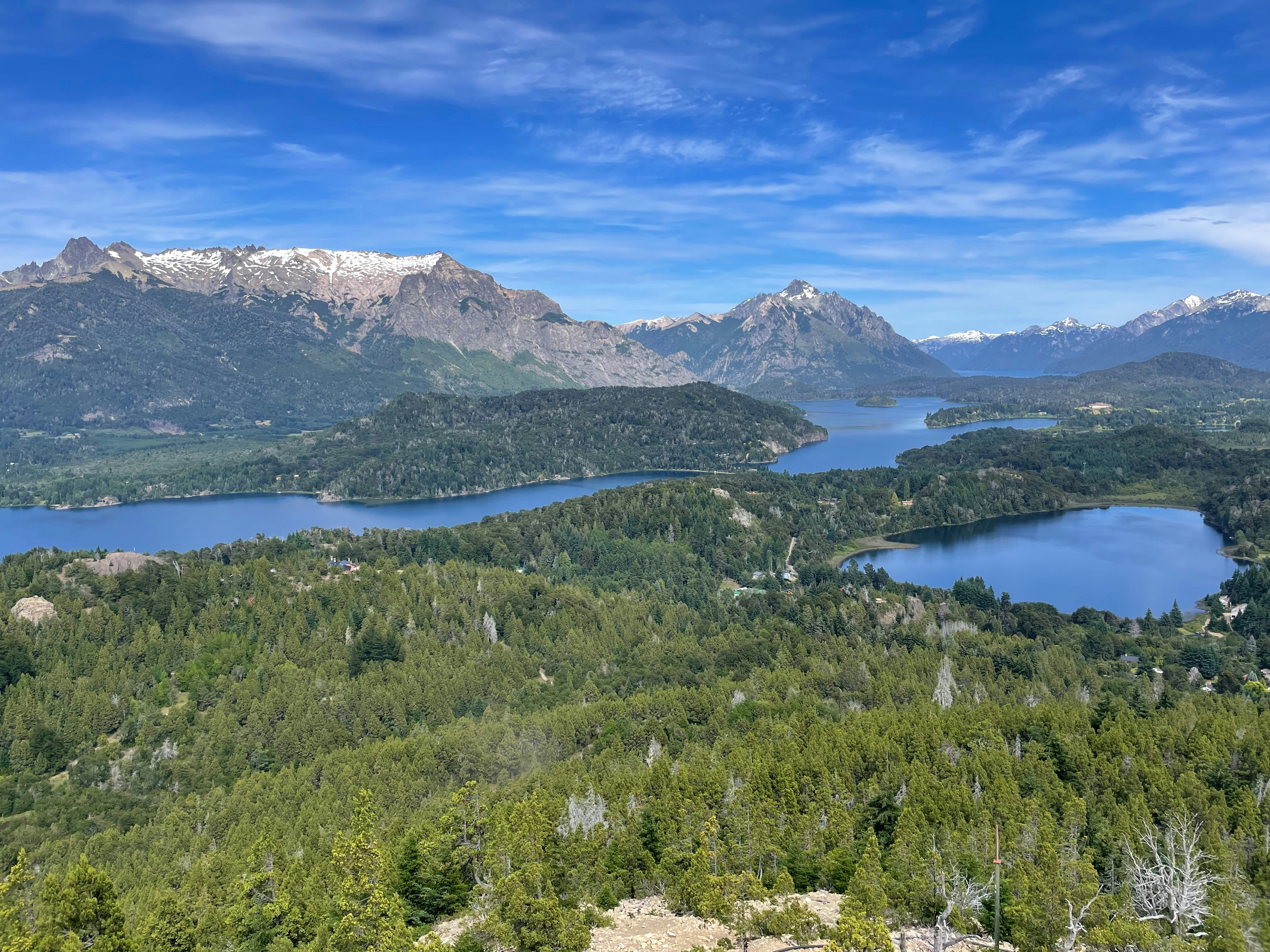 Lush green forest surrounds blue lakes with snowy mountains.