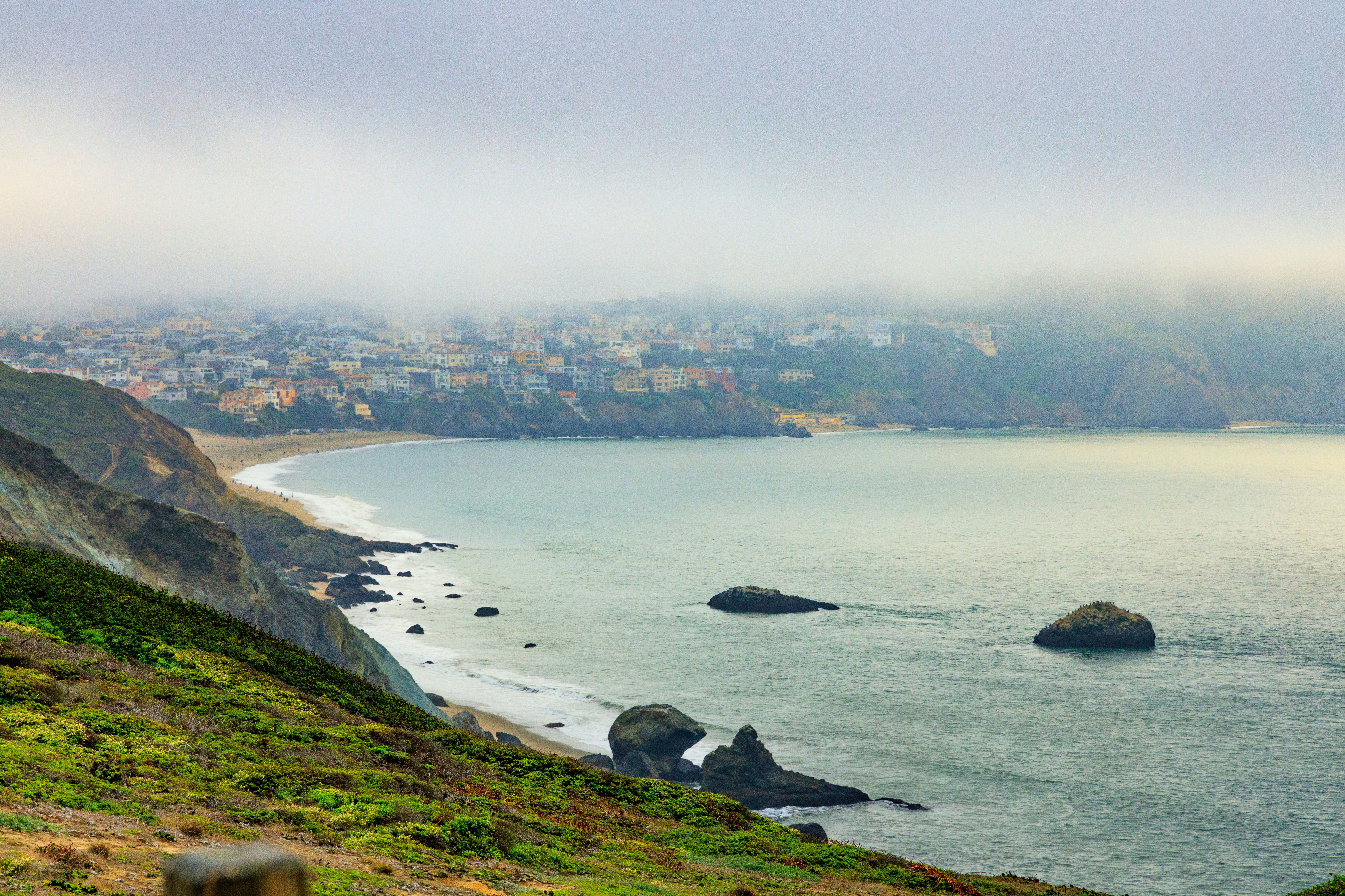 Foggy coastal town with ocean rocks and green hills.