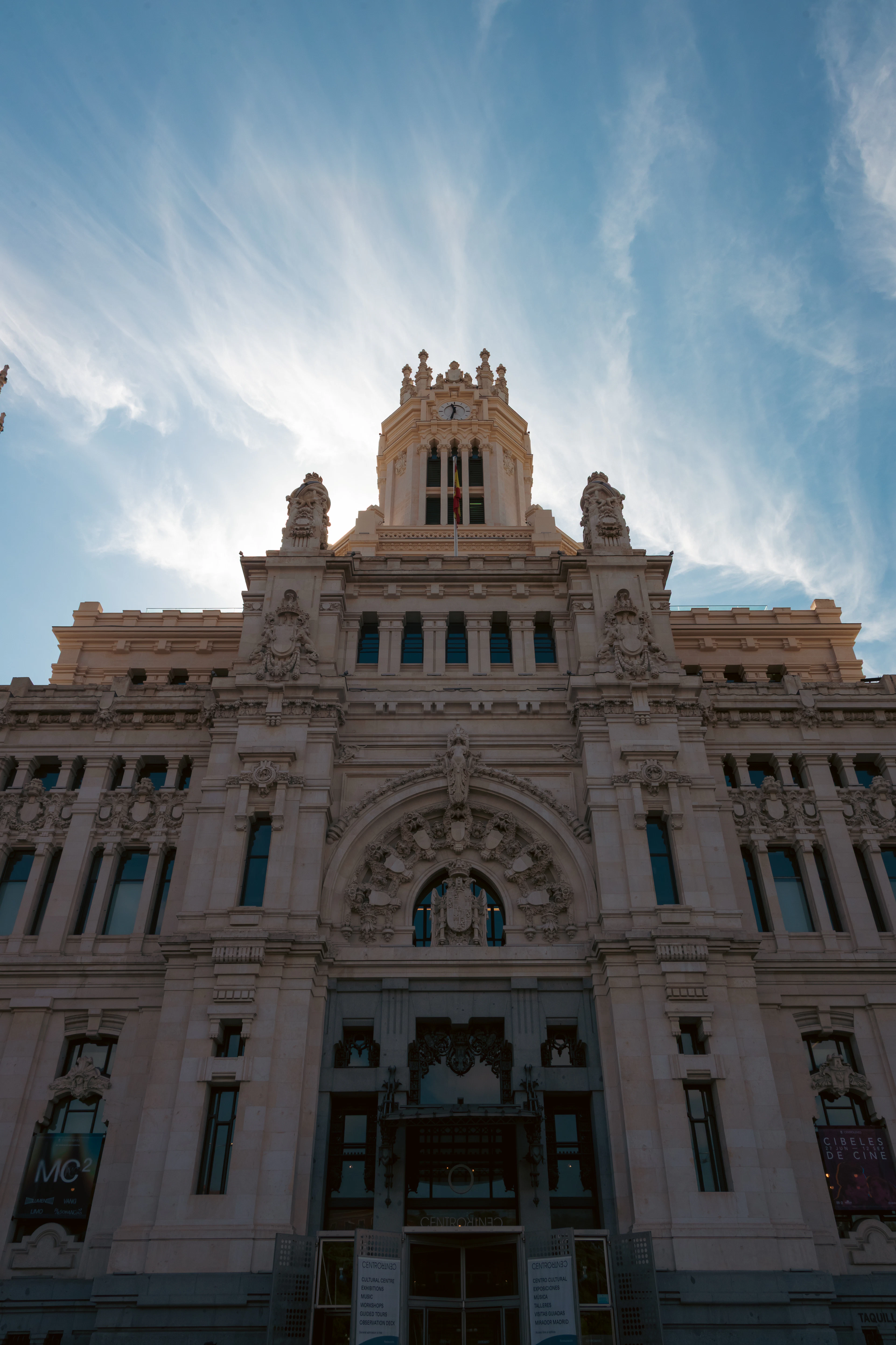 The Palacio de Cibeles seen from the pavement directly below, its white baroque facade and clock tower rising into a deep blue sky streaked with cirrus cloud.