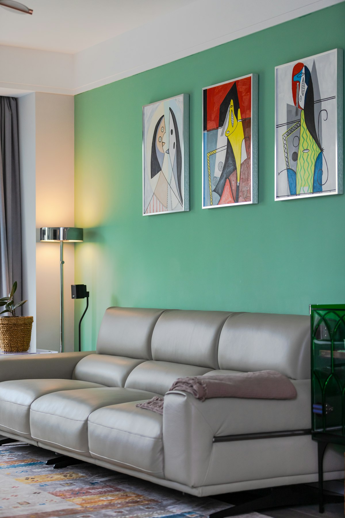 Living con un sillón de cuero gris claro frente a una pared pintada de verde, con tres cuadros de arte abstracto en marcos plateados colgados en fila, una lámpara de pie cromada encendida y una planta en canasto de fibra