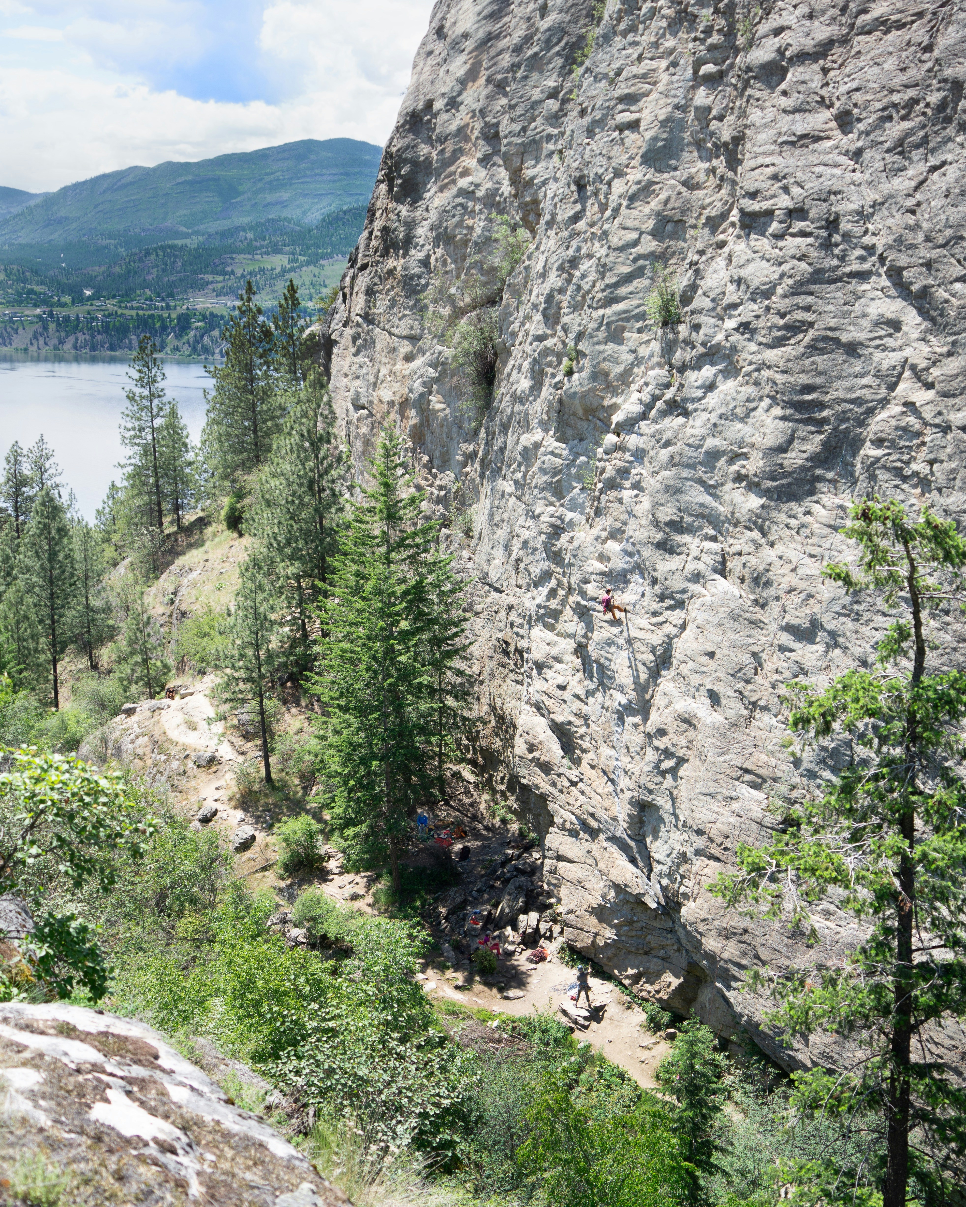Rock climbers ascend a sheer cliff face near a lake.