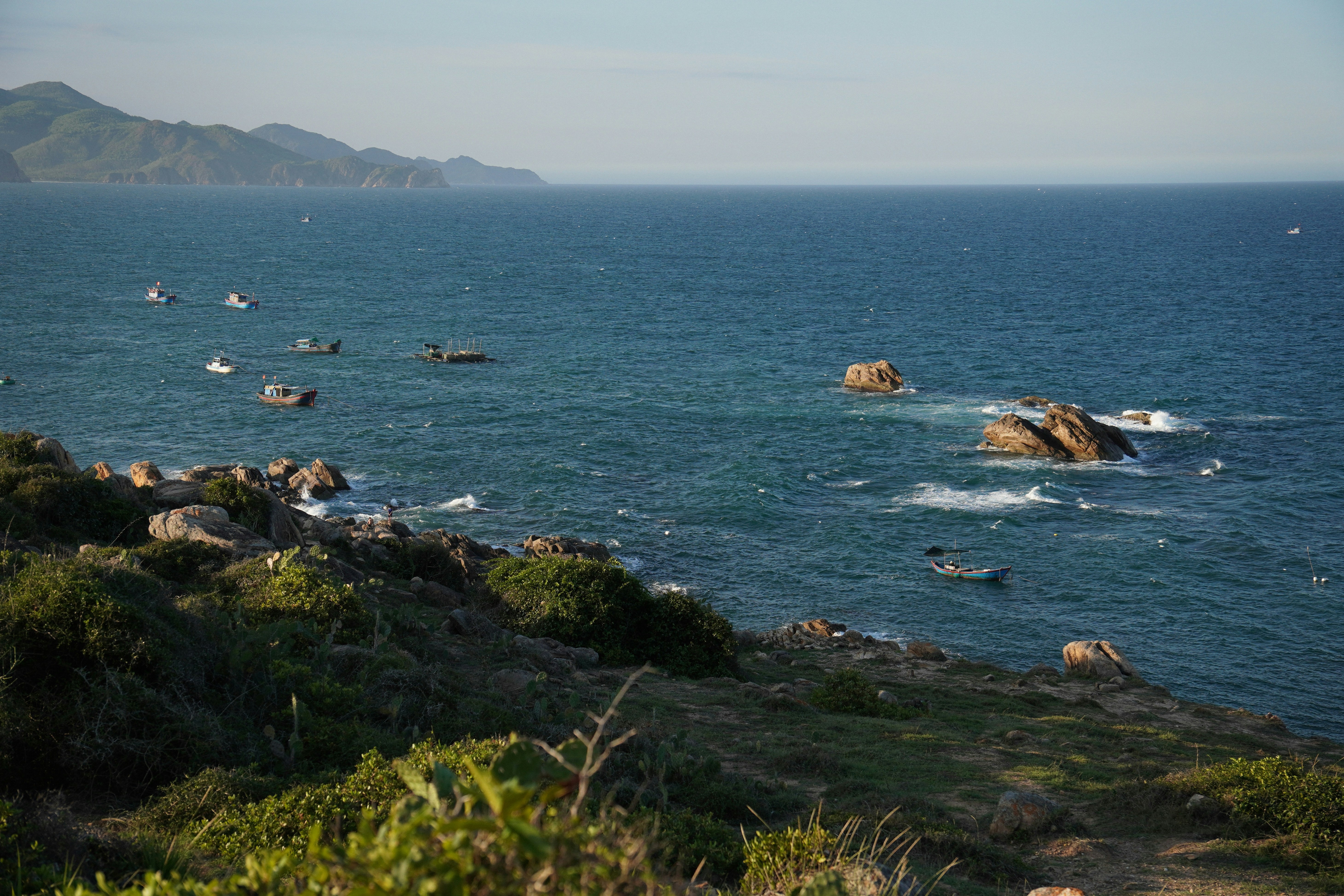 Boats dot the ocean near a rocky, vegetated coastline.