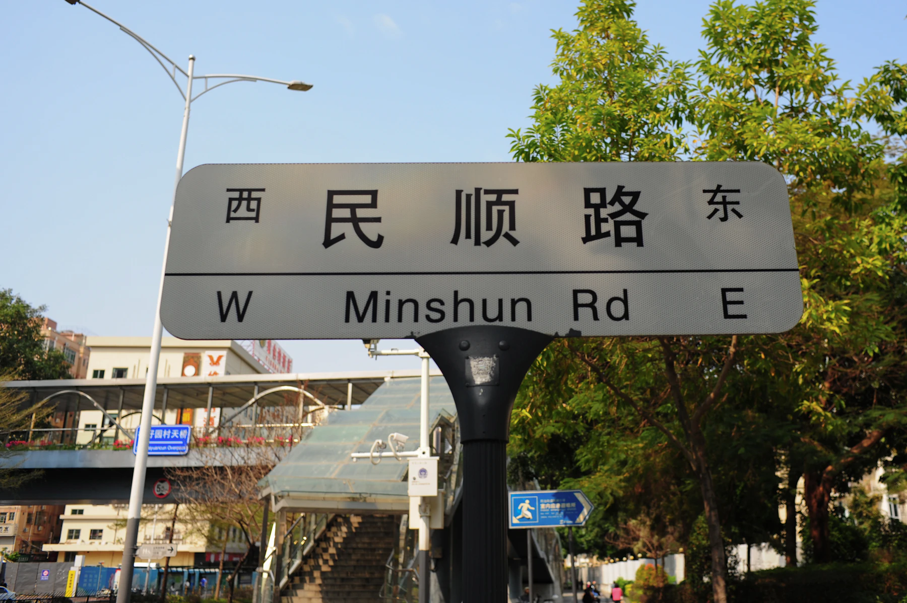A gray road sign against a blue sky, four Chinese characters on its top line and the road's name in pinyin on the line below