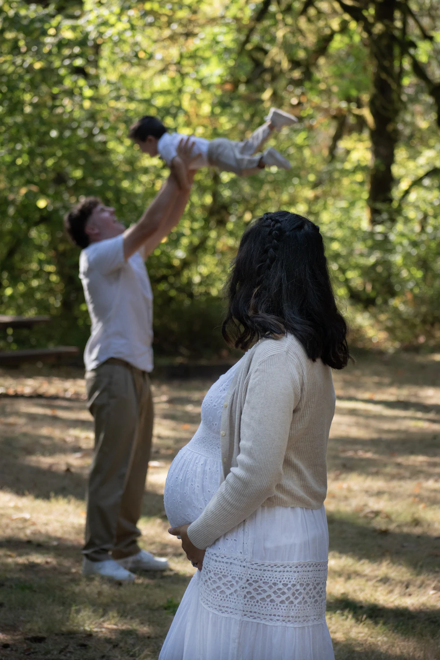 Father tossing his child in the air in the woods as the expecting mother watches