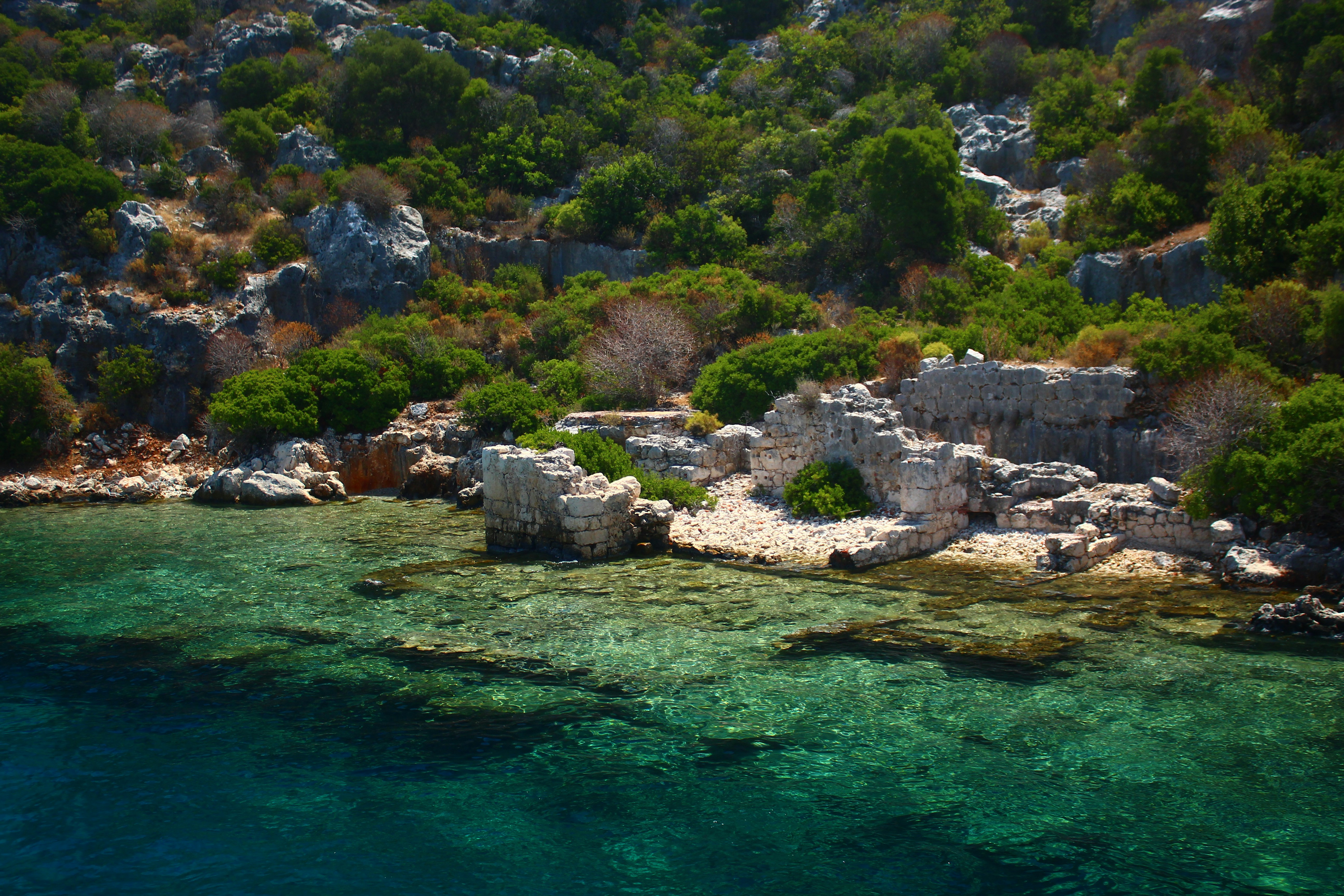 Ancient ruins on a rocky coastline beside clear turquoise water.