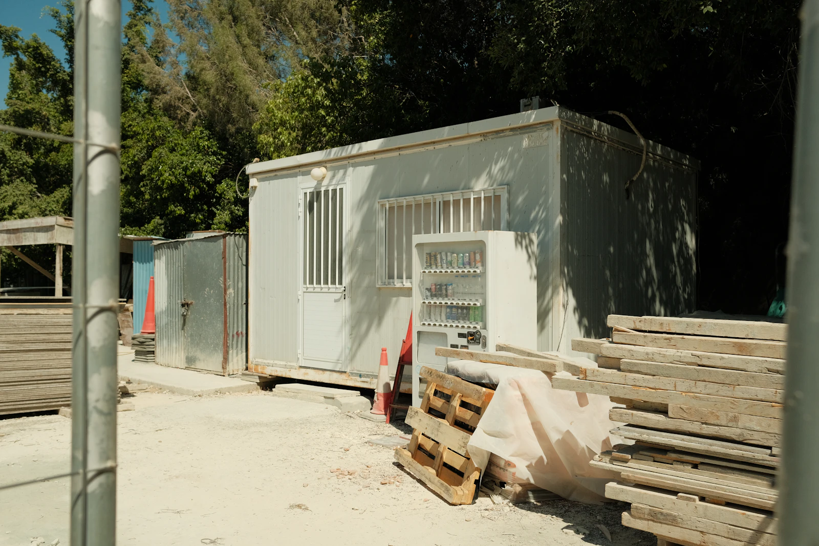 A transportable site office behind temporary fencing, with a drinks machine, stacked formwork timber and a traffic cone beside it