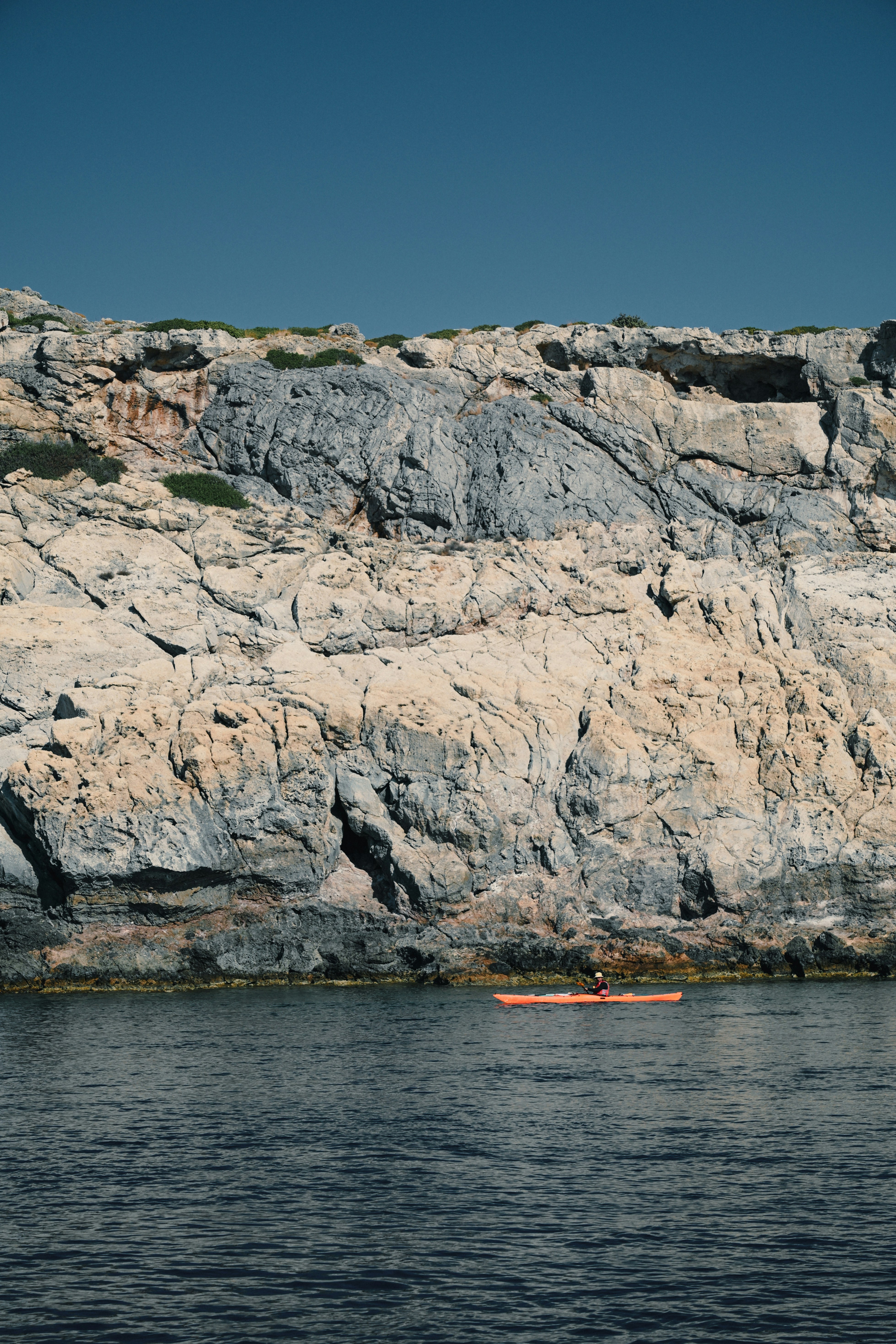 Person kayaking near rocky coastline under clear blue sky.