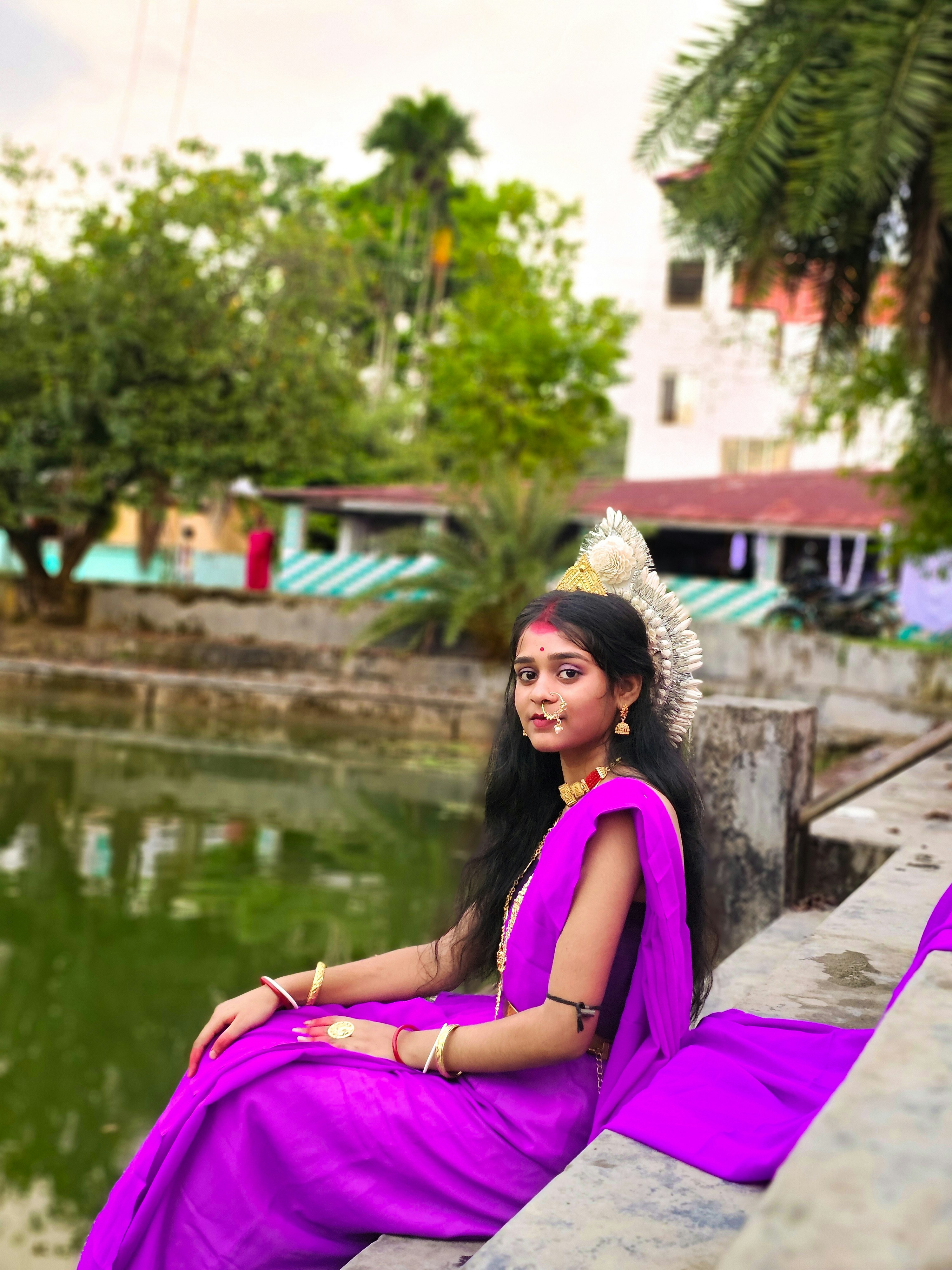 Young woman in a purple sari sits by water.