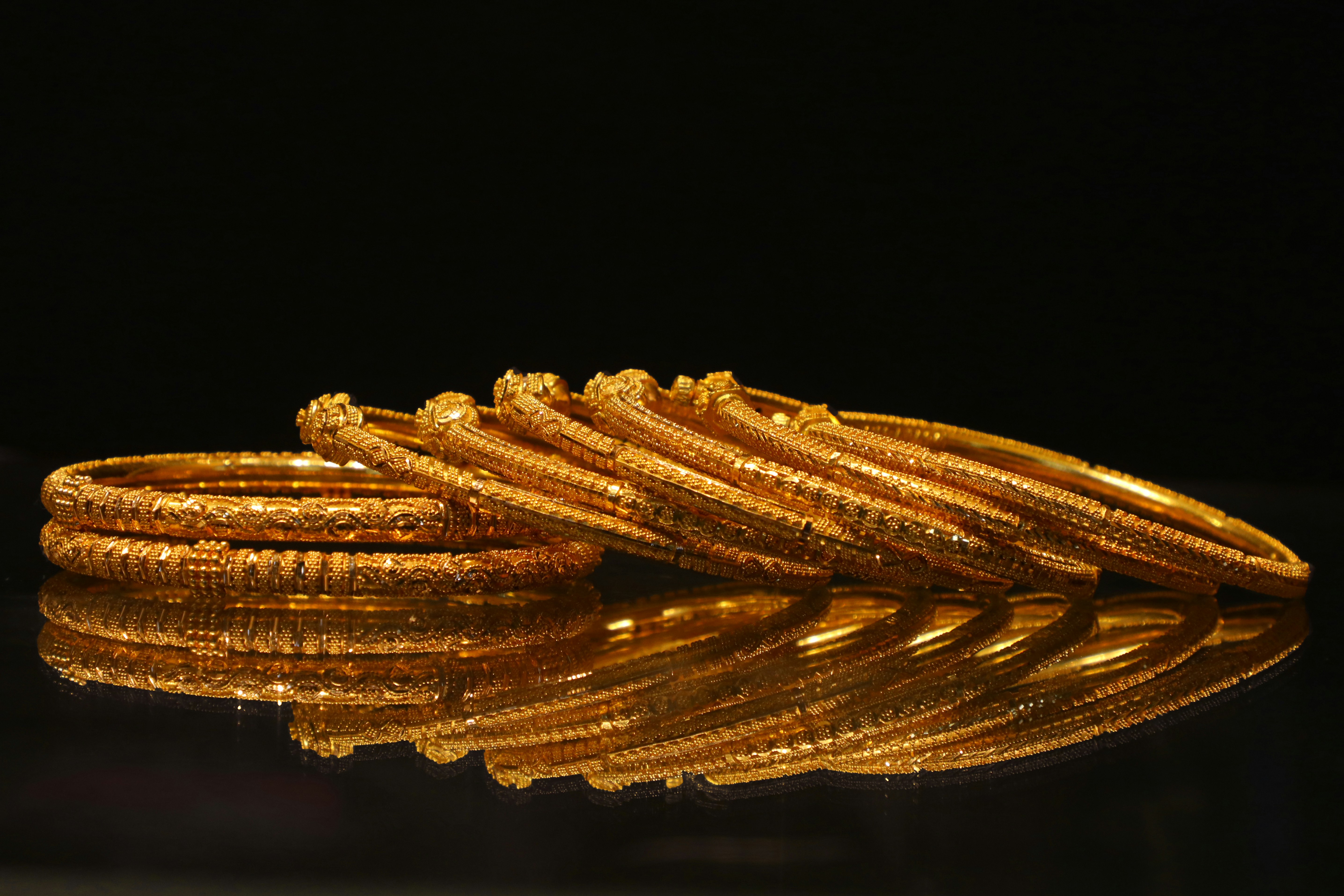 A set of intricately designed gold bangles with ornate patterns, placed on a reflective black surface, showcasing their rich shine, elegance, and traditional craftsmanship.
