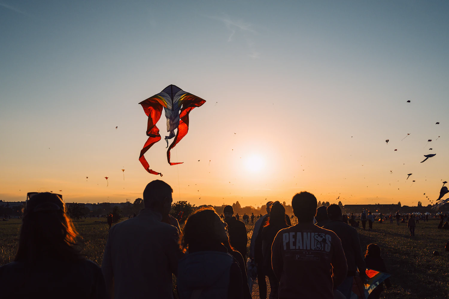 A red hawk kite flying at sunset above a crowd watching from the field