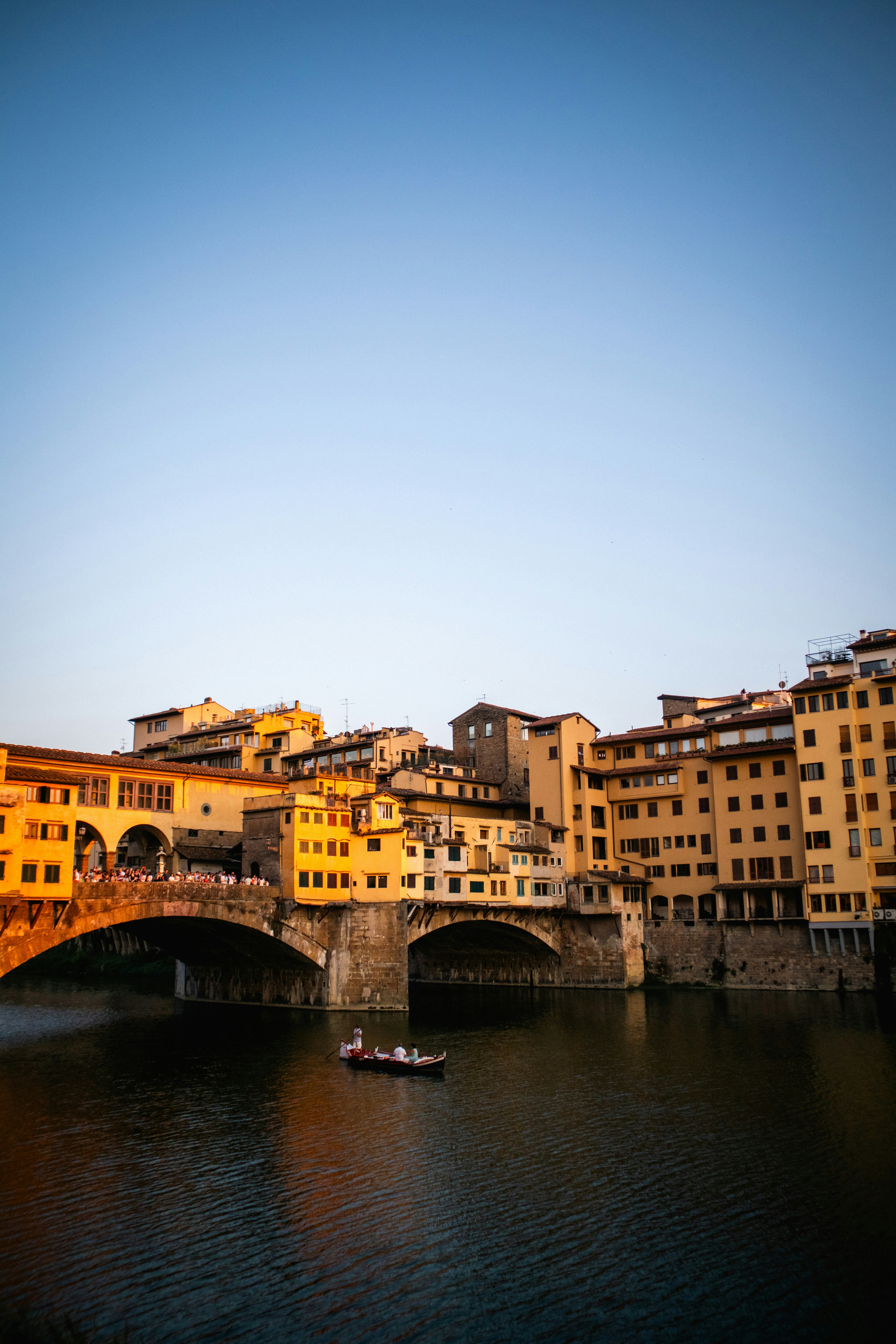 Boat on river beneath ponte vecchio bridge at sunset.