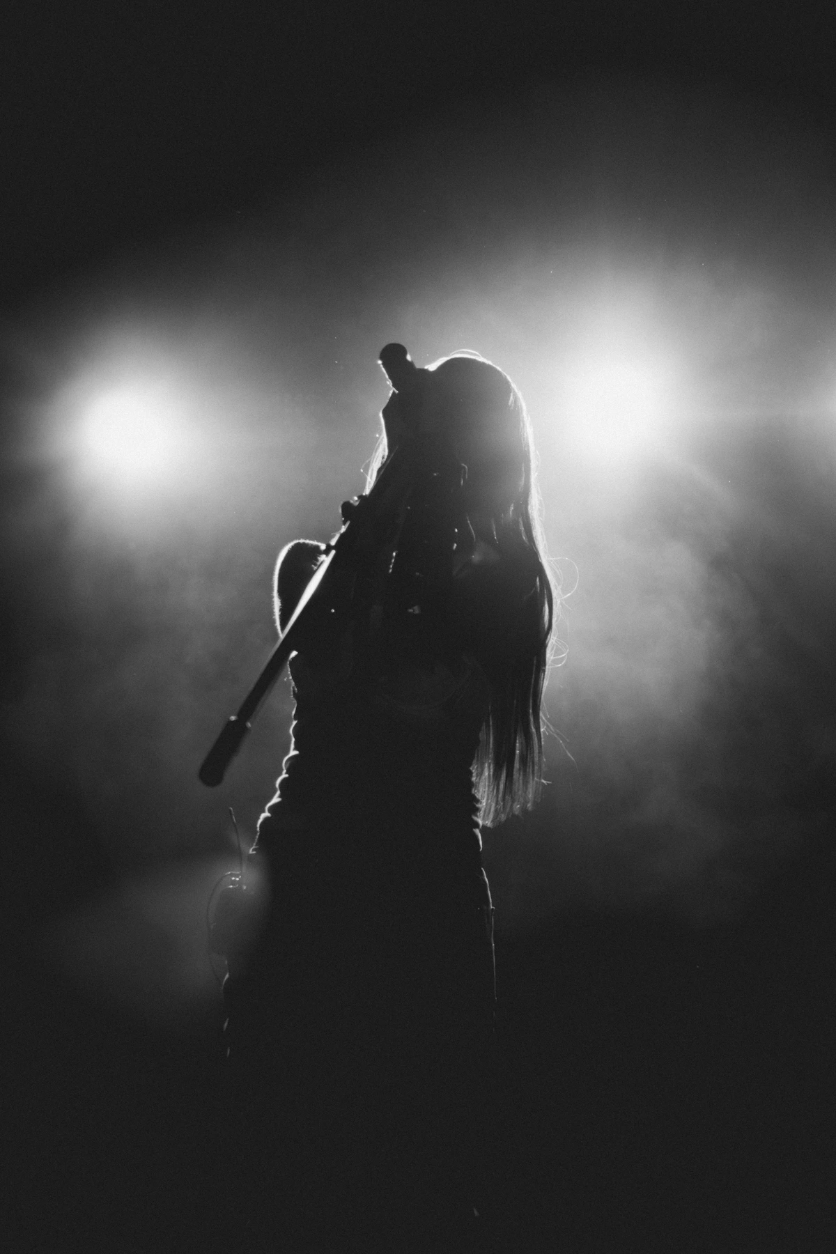 A monochrome silhouette of a vocalist singing, gripping a microphone stand, against the bright backlight of the stage.