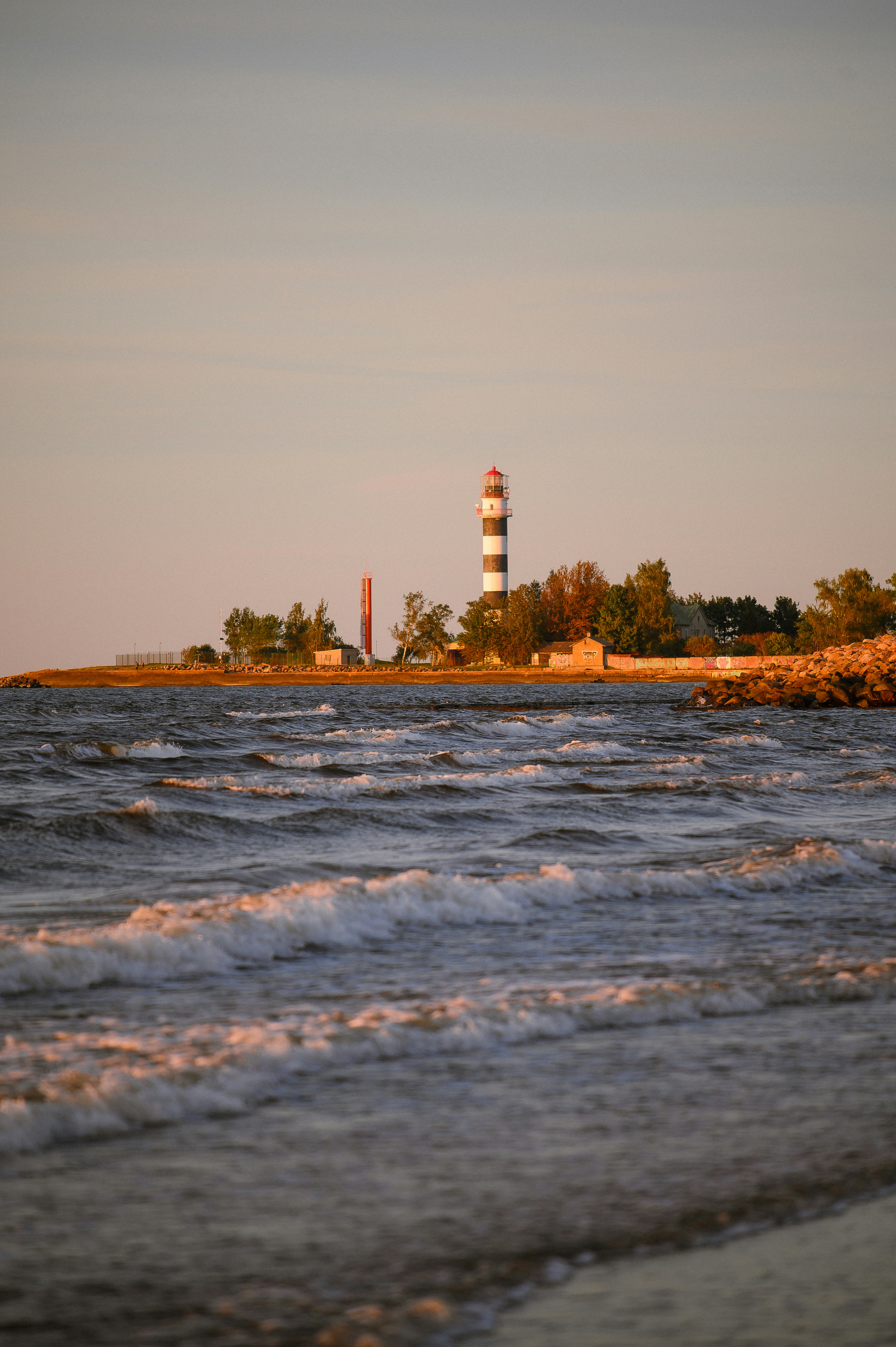 Lighthouse on a rocky shore with waves at sunset.