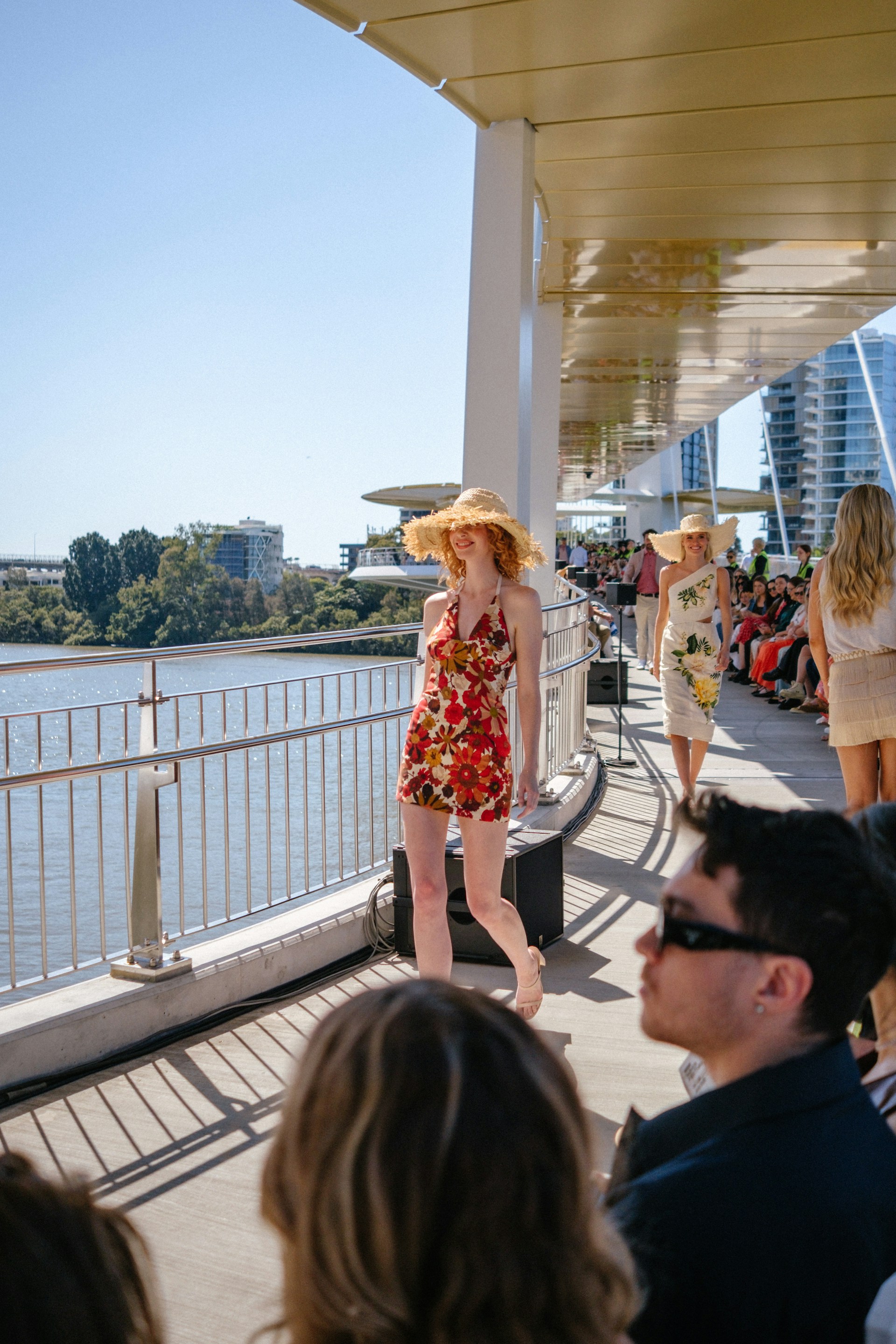 Models walk on a sunny outdoor runway by the water.