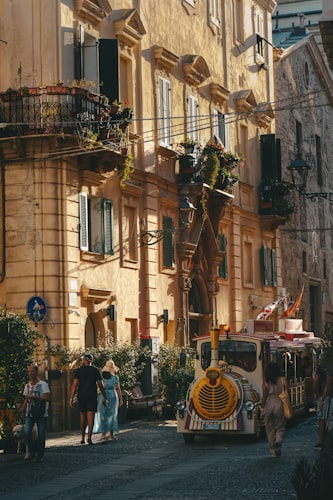 Kopfsteinpflasterstraße in der Altstadt von Alghero mit kunstvollen Steingebäuden und einem vorbeifahrenden Touristen-Zug