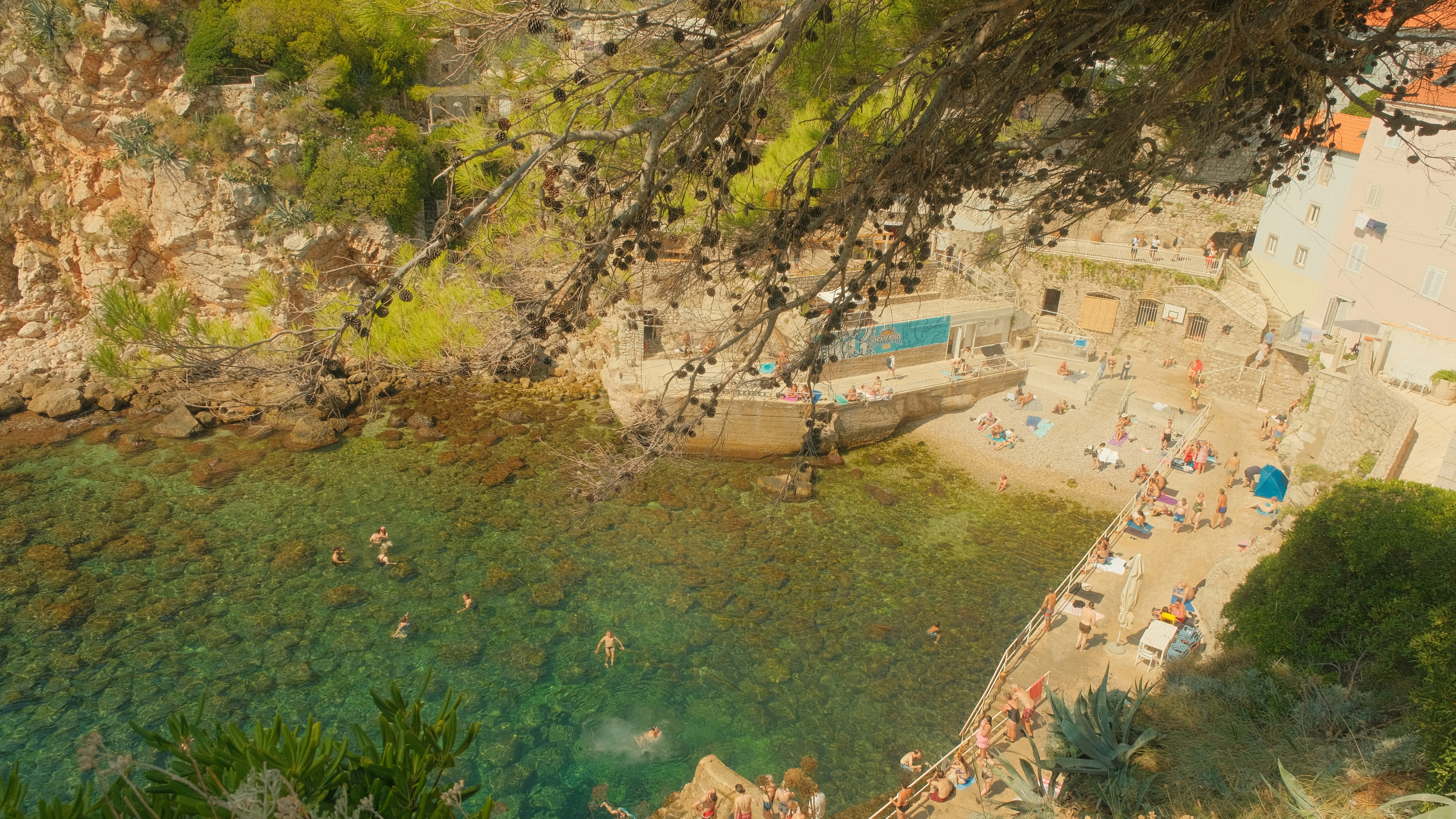 A vibrant, high-angle view of the picturesque Šulić Beach, a beautiful cove nestled just outside the walls of Dubrovnik, Croatia. Swimmers enjoy the crystal-clear turquoise water while others relax on the sun-drenched shore, creating a perfect summer scene.