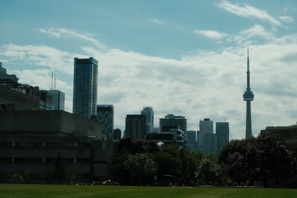 The image depicts a cityscape with a tall, gray building on the left side and a smaller, white building on the right side. The buildings are surrounded by lush green trees, creating a contrast between the urban architecture and nature's presence. In the distance, there is a large tower that stands out against the sky.