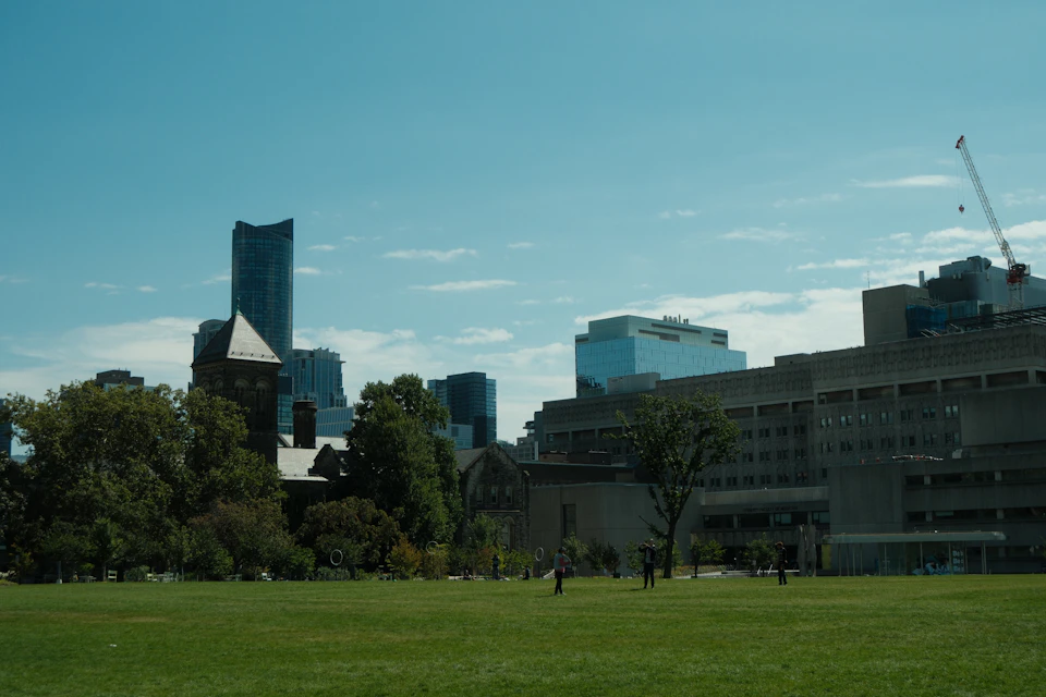 The image depicts a cityscape with a large, modern glass building on the left side and a smaller, older brick building on the right side. The buildings are situated amidst lush green trees, creating a contrast between the old and new architectural styles. In the foreground, there is a grassy field where several people can be seen walking or standing, adding a sense of scale to the scene.