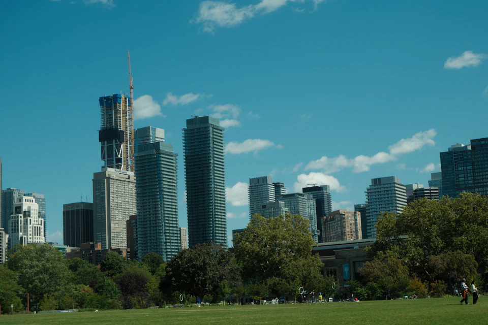 The image depicts a bustling cityscape with tall buildings, including a prominent skyscraper on the left side and several other structures of varying heights in the background. The sky above is a clear blue, dotted with fluffy white clouds that add depth to the scene. In the foreground, there's a large park filled with lush green grass, providing a contrast to the urban environment. Several people can be seen walking around the park or sitting on benches scattered throughout the area.