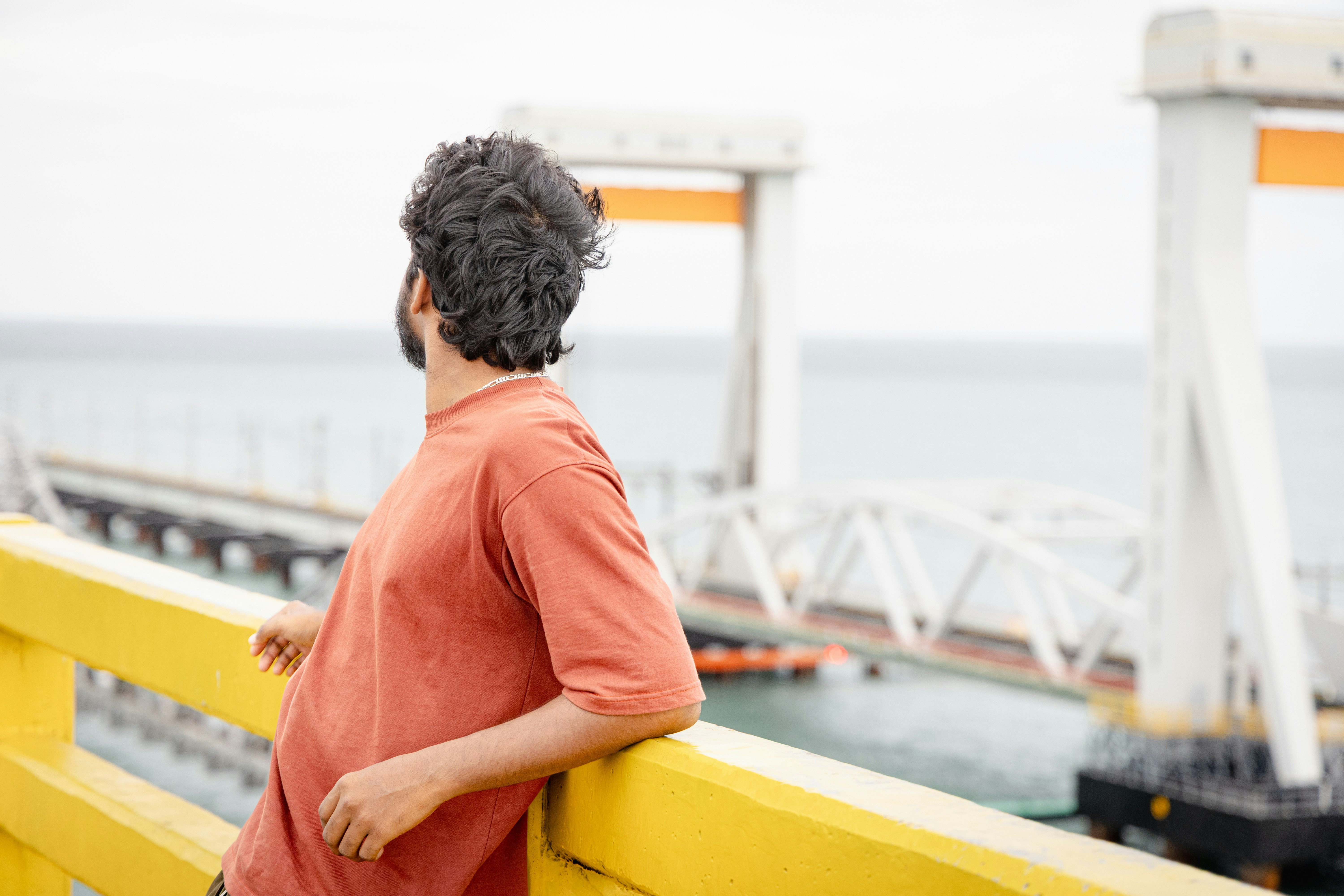 Hombre apoyado en una barandilla amarilla con vistas al océano y al puente.