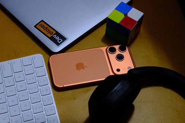 Laptop, phone, keyboard, headphones, and puzzle cube on desk.