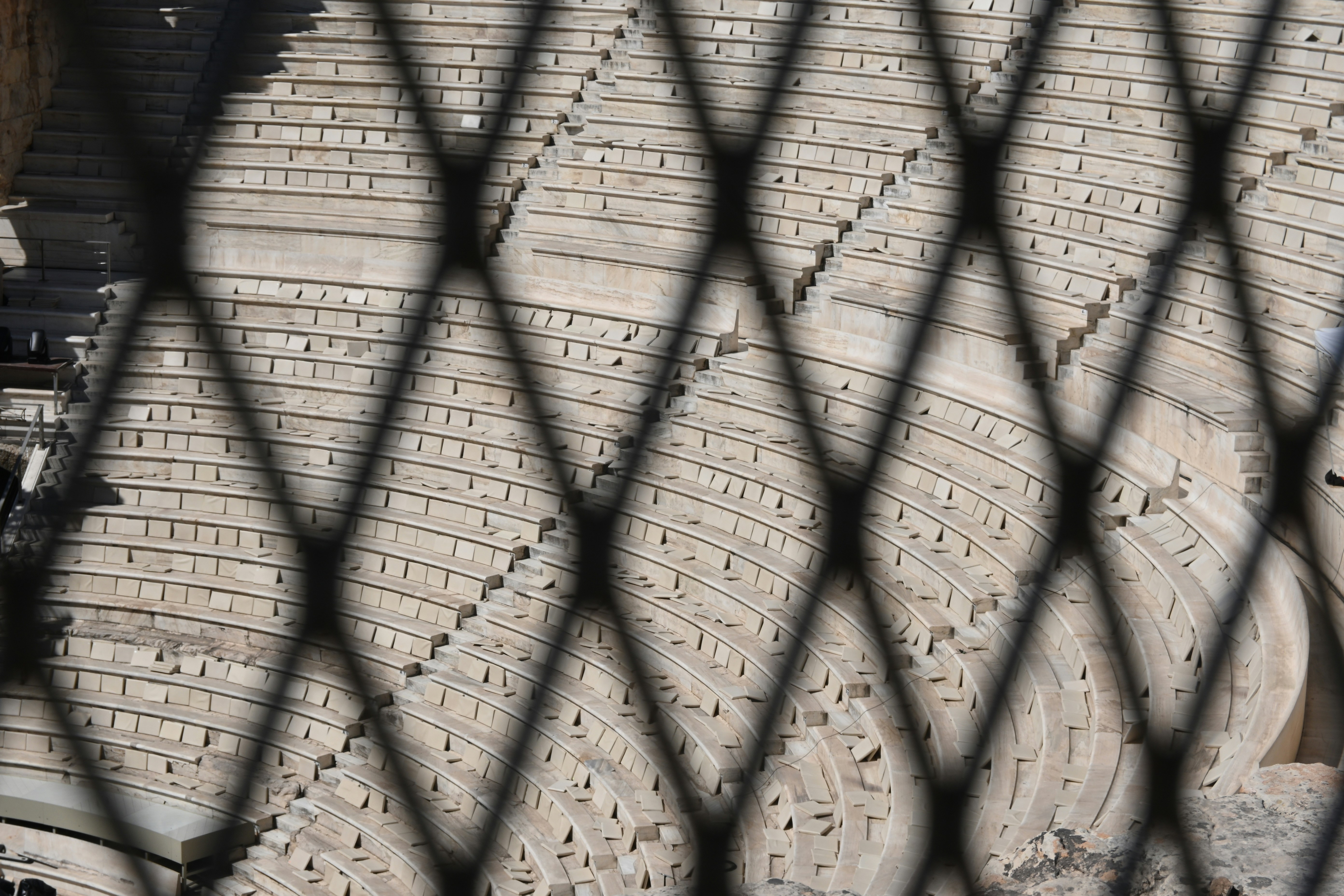 Stone amphitheater seating viewed through a diamond pattern fence.